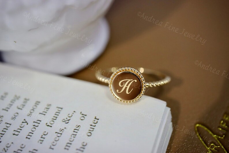 May include: A gold ring with a round, beaded band and a circular top featuring the letter "H" engraved in a cursive font.