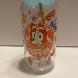 May include: A clear glass tumbler with a blue lid and a rainbow straw. The tumbler has a cartoon image of a dog with the words "Bluey Coffee" on it. The tumbler is decorated with various cartoon images, including a sun, flowers, sunglasses, and a watermelon.