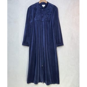 Puede incluir: Una bata larga de terciopelo azul marino con mangas largas y cremallera frontal. La bata tiene un cuello redondo y un panel decorativo en el pecho con detalles de botones. La bata está colgada en una percha de madera sobre un fondo neutro.