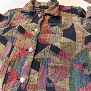 Puede incluir: Chaqueta multicolor con un patrón geométrico. La chaqueta tiene cuello, tres botones decorativos y el texto "TO DIE FOR" en el cuello. La paleta de colores incluye rojo, verde, morado y negro.