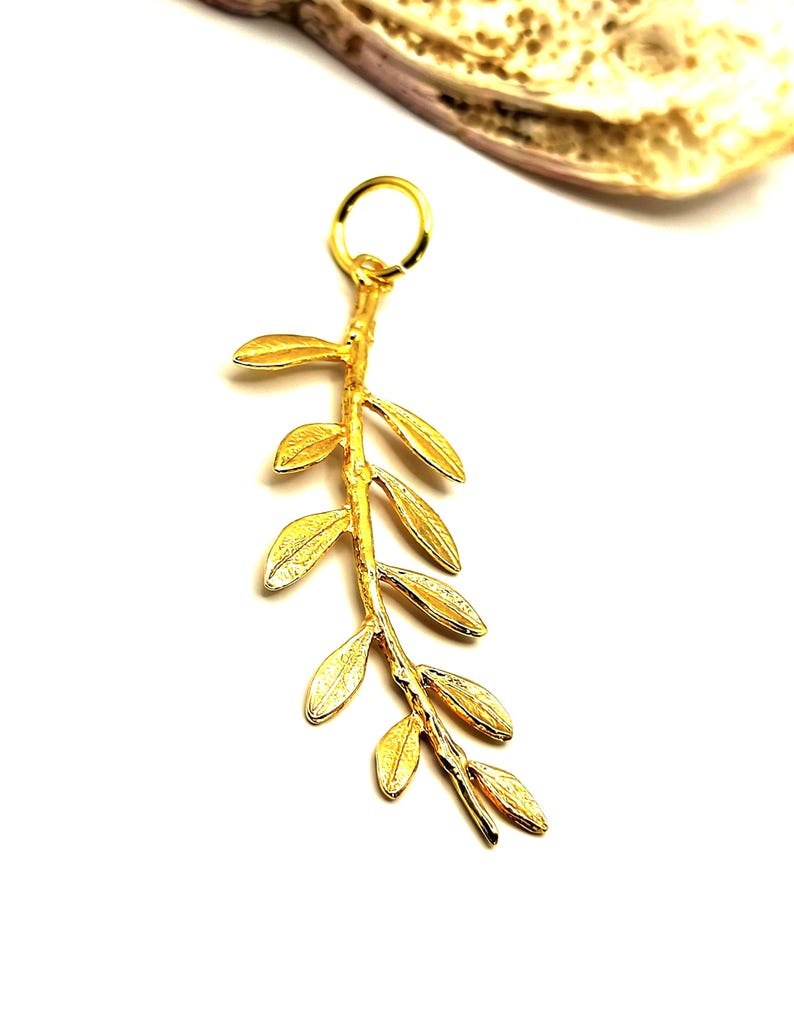 May include: A gold-colored pendant charm in the shape of a branch with leaves. The charm is made of metal and has a loop at the top for attaching to a necklace or bracelet.