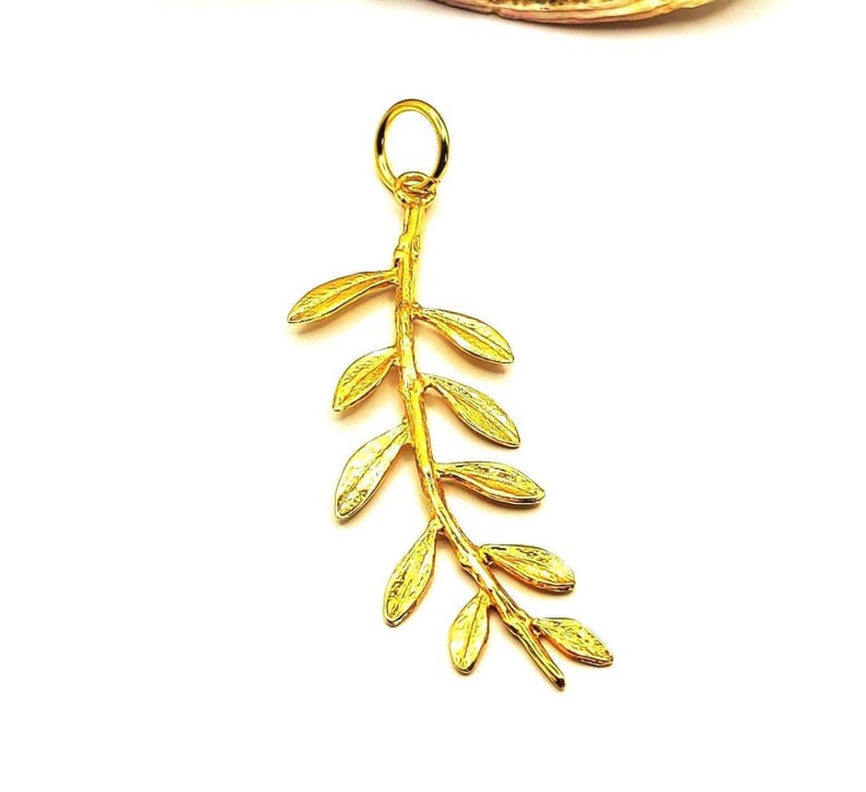 May include: A gold-colored pendant in the shape of a branch with leaves. The pendant is made of metal and has a smooth, polished finish.
