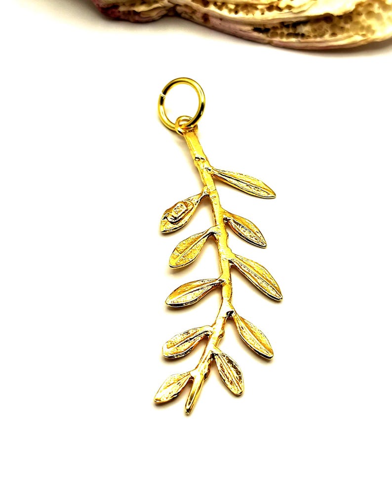 May include: A gold-colored pendant in the shape of an olive branch with leaves. The pendant is on a white background.