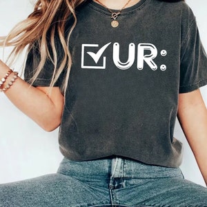 May include: A dark gray t-shirt with a white checkmark in a box and the text "UR:" in white.
