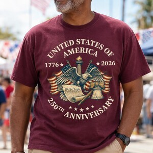 May include: Burgundy t-shirt with a graphic commemorating the 250th anniversary of the United States of America. The design includes an eagle, flags, a scroll, and the text "United States of America 1776-2026 250th Anniversary."