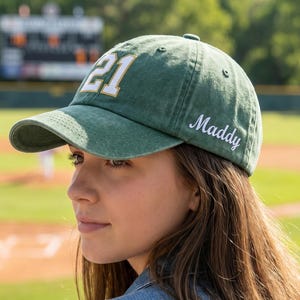 May include: A green baseball cap with a distressed finish featuring the number 21 embroidered in white with a gold outline on the front and the name Maddy embroidered in white cursive script on the side panel above the brim.