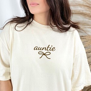 May include: Cream-colored t-shirt with the word "auntie" in a brown script font, accompanied by a bow graphic. The shirt has short sleeves and a relaxed fit, suitable for casual wear.