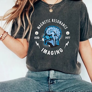 May include: A dark grey t-shirt with a vintage-style graphic design featuring a blue brain inside a head silhouette. The text "Magnetic Resonance Imaging" is written in a circle around the head, with "Estd 1977" below.