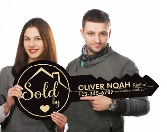Può includere: Un cartello a forma di chiave nero e dorato con il testo "Venduto da" e un cuore. Il cartello è tenuto da due persone, un uomo e una donna. L'uomo sta indicando il testo "OLIVER NOAH Agente immobiliare 123-345-6789 www.olivernoah.com".