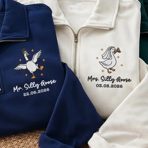 May include: Three quarter-zip sweatshirts in navy blue, cream, and dark green. Each features an embroidered goose graphic, text reading Mr. Silly Goose or Mrs. Silly Goose, and a specific date. The sweatshirts have long sleeves and metal zippers.