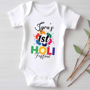May include: White baby onesie with a colourful design that says "Syra's 1st Holi Festival".