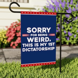 May include: A navy blue garden flag with white text that reads "SORRY FOR BEING WEIRD, THIS IS MY 1ST DICTATORSHIP." The flag has red and white star accents at the top and bottom. The flag is displayed on a black metal stand in a grassy yard.