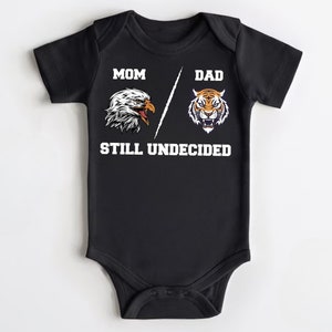 May include: Black baby growsuit with a white eagle on the left side and a tiger on the right side. The text "MOM" is above the eagle, "DAD" is above the tiger, and "STILL UNDECIDED" is below the animals.