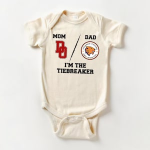 May include: A white baby onesie with the text "Mom / Dad I'm the Tiebreaker" printed on it. The onesie also features a red "D" and a circular orange logo with a tiger head and the text "Orange Tigers Est. MMXV"