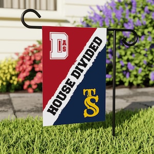 May include: A red and blue garden flag with a white diagonal line. The text "HOUSE DIVIDED" is written on the white line. The top half of the flag features a white "D" with the letters "AS" inside. The bottom half of the flag features a yellow "S" with a "T" above it.