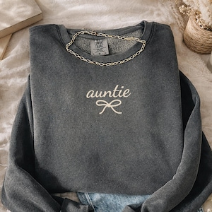 May include: A gray sweatshirt with the word "auntie" embroidered in white script, along with a bow design. A gold chain necklace rests on the collar. The sweatshirt is paired with denim shorts.