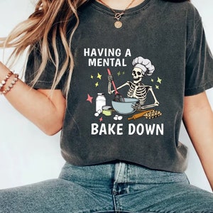 May include: A gray t-shirt with a skeleton wearing a chef's hat and holding a whisk. The text on the shirt reads "Having a mental bake down."
