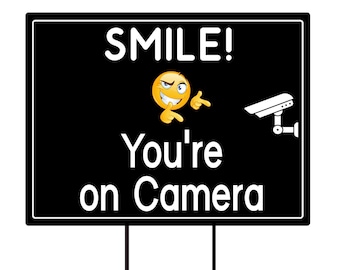 May include: Black and white sign with the text "SMILE! You're on Camera" in white. A yellow smiley face with a wink and pointing fingers is above the text. A black and white security camera icon is on the right side of the sign.