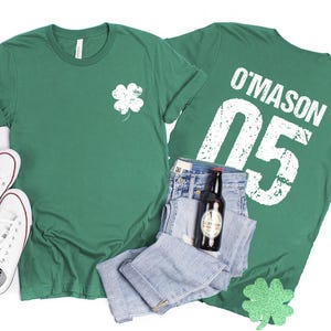 May include: Two green t-shirts, one with a white shamrock and the other with "O'MASON 05" printed on the back. The shirts are paired with blue jeans, a bottle of beer, and white sneakers. Decorative shamrock cutouts are also included.