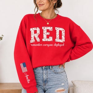 May include: A bright red crewneck sweatshirt featuring the word RED in large white block letters with a floral pattern. Below the word, the text reads remember everyone deployed. The right sleeve displays a small American flag and the name Amelia.