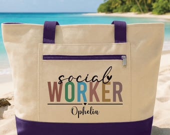 May include: A canvas tote bag with a cream-colored body, purple accents, and a zippered pocket. The bag features the words "social WORKER" in colorful letters, with the name "Ophelia" below. The bag is on a sandy beach with a turquoise ocean in the background.