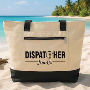 May include: A beige canvas tote bag with black accents, a zippered pocket, and black handles. The bag features the text "DISPATCHER" above "Amelia" in black lettering. The bag is on a sandy beach with a tropical background.
