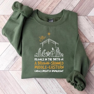 May include: Green sweatshirt with a white graphic of a nativity scene and the text "Rejoice in the birth of a brown-skinned Middle-Eastern undocumented immigrant".