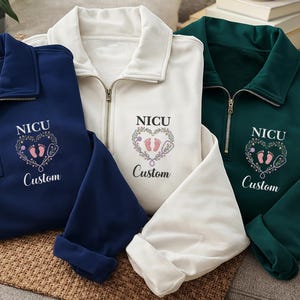 May include: Three quarter-zip sweatshirts in navy, white, and dark green. Each has "NICU" above a heart-shaped design with baby footprints and a stethoscope, and the word "Custom" below.