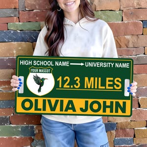 May include: A green and yellow sign with the text "HIGH SCHOOL NAME -> UNIVERSITY NAME", "YOUR MASCOT", "12.3 miles", and "OLIVIA JOHN". The sign features a hummingbird graphic. The sign is held up in front of a brick wall.