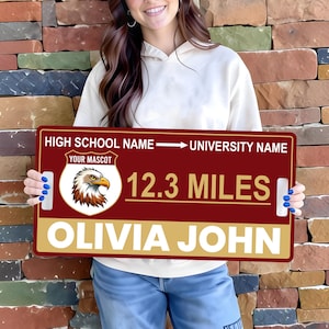 May include: A maroon and gold sign with the text "HIGH SCHOOL NAME -> UNIVERSITY NAME", "YOUR MASCOT", and "12.3 MILES". The sign also includes the name "OLIVIA JOHN" and an eagle graphic. The sign is held by a person.