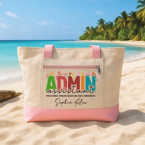 May include: A beige tote bag with pink accents, featuring the text "ADMIN ASSISTANT" in colorful letters, with floral embellishments. The bag has a zippered pocket and pink handles, perfect for carrying essentials.