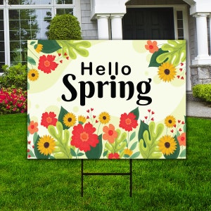 May include: A white yard sign with a green, yellow, and red floral design. The sign says "Hello Spring" in black text.