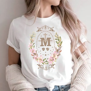 Op de afbeelding: Wit T-shirt met een bloemenkransontwerp met een bruine "M" met een kruis en twee harten. De tekst luidt "O Maria, ontvangen zonder zonde, bid voor ons die onze toevlucht tot u nemen."