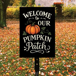 May include: A black sign with the words "WELCOME to OUR PUMPKIN Patch" in white script. An orange pumpkin with green vines is in the center. The sign is on a black metal post, set in a grassy area covered in fallen leaves.