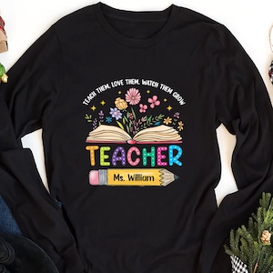 May include: Black long-sleeve shirt with a colorful graphic of an open book with flowers and the word "TEACHER" in rainbow letters. A yellow pencil with a pink eraser is below the book with the text "Ms. William" on it.