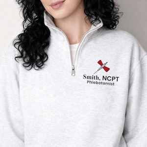 May include: Light gray quarter-zip sweatshirt with a white collar and a silver zipper. The sweatshirt features a red and black medical symbol and the text "Smith, NCPT Phlebotomist" in black.