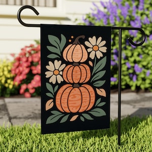 May include: A black garden flag featuring a stacked pumpkin design with daisies and leaves. The pumpkins are orange with brown stems, and the flowers are white with yellow centers. The flag is displayed on a black metal stand in a grassy yard.