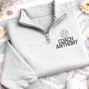 May include: A light grey baseball-themed pullover sweatshirt with a quarter-zip collar. The sweatshirt features a baseball graphic and the text "COACH ANTHONY" in black. The sleeves are long, and the cuffs and collar are ribbed.