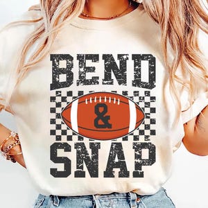 May include: Cream-colored t-shirt with the words "BEND & SNAP" in distressed black lettering. A football graphic is centered, with a checkered pattern behind it. The football is orange and white.