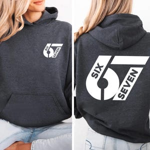 May include: Charcoal gray hoodie with a front pocket and a hood. The front and back of the hoodie feature a white graphic design with the text "SIX SEVEN". The hoodie is made of a soft material.