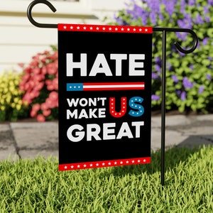 May include: Black garden flag with white text that reads "Hate Won't Make Us Great" with a red, white, and blue "US" in the middle. The flag is attached to a black metal garden stake.