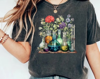 Può includere: Una maglietta grigio scuro con un'illustrazione colorata di fiori e bottiglie di vetro riempite di liquidi di diversi colori. Le bottiglie sono disposte su uno scaffale di legno.
