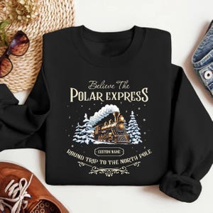 May include: Black sweatshirt with a vintage illustration of a train with the text "Believe the Polar Express Round Trip to the North Pole" and a space for a custom name.