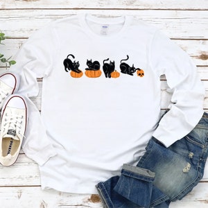 May include: White long-sleeved t-shirt with a black cat design. The cat is holding a pumpkin and a skull.