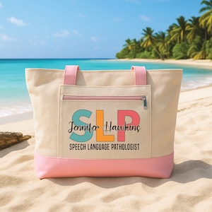 May include: Beige tote bag with pink detailing, displaying the letters "SLP" in teal, orange, and pink, plus the name "Jennifer Hawkins" and the words "Speech Language Pathologist". The bag has a zipped pocket and pink handles.
