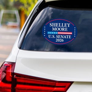 May include: An oval sticker on a car window. The dark blue sticker has white text: "SHELLEY MOORE" and "U.S. SENATE 2026." It features a red and white stripe design and white stars around the edge.