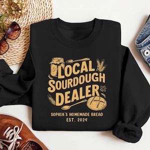 May include: Black sweatshirt with a gold graphic design that says "Local Sourdough Dealer" with a jar of sourdough starter, a spoon, a loaf of bread, and wheat stalks. The text below the graphic says "Sophia's Homemade Bread Est. 2024".