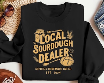 May include: Black sweatshirt with a gold graphic design that says "Local Sourdough Dealer" with a jar of sourdough starter, a spoon, a loaf of bread, and wheat stalks. The text below the graphic says "Sophia's Homemade Bread Est. 2024".