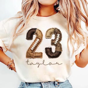 May include: A cream-coloured short-sleeve t-shirt featuring the number 23 designed with a brown and tan football-themed texture, including white laces. Below the number, the name Taylor is printed in a brown cursive script font across the chest.