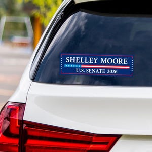 May include: A rectangular bumper sticker with the text "SHELLEY MOORE U.S. SENATE 2026" in white letters on a blue background. The sticker has a red and white stripe design and a row of white stars. The sticker is affixed to the rear window of a white car.